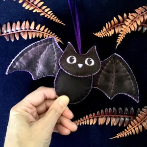 May include: A black felt bat decoration with purple stitching and a purple ribbon. The bat features white eyes and a small white triangle nose. The wings are a darker shade of black. Dried brown fern leaves are in the background.