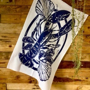 May include: A white tea towel with a blue illustration of a lobster, seashells, and mussels. The design is in a circular frame.