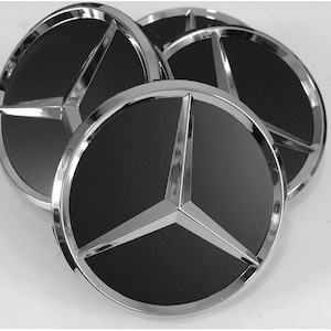 May include: Four black and silver car wheel center caps. Each cap features a three-pointed star emblem within a circular design. The caps have a shiny, chrome-like finish around the edge.