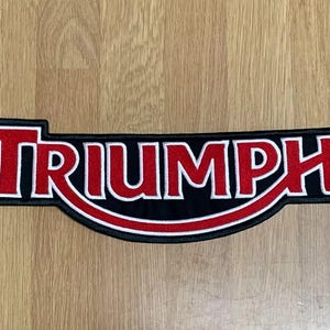 Peut inclure: Écusson Triumph noir et rouge avec bordure blanche. Le mot "TRIUMPH" est en lettres rouges grasses avec un contour blanc, sur fond noir. L'écusson a un bord inférieur incurvé.