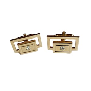 May include: A pair of gold cufflinks with a square design and a single clear rhinestone in the center of each cufflink.