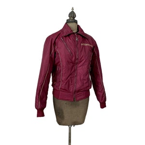 May include: A maroon quilted jacket with a zipper closure and white piping. The jacket has a small pocket on the left chest with the word "INTERSTATE" embroidered in gold.