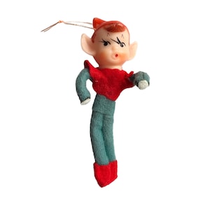Puede incluir: Adorno navideño vintage de elfo. Presenta una cara de plástico, pelo y gorro rojos, y una parte superior de fieltro rojo. El elfo tiene brazos y piernas de fieltro verde azulado, botas de fieltro rojo y una mano de fieltro blanco. Una cuerda está unida para colgar.