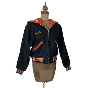 May include: Black vintage zip-up jacket with orange accents. The jacket features an orange hood lining, pocket trim, and striped cuffs and hem. The name "Tricia" is embroidered on the chest. A classic varsity style jacket.