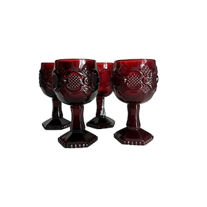 May include: Four ruby red wine glasses with ornate, textured designs. The glasses have a classic goblet shape with a detailed pattern on the bowl and stem. The glasses are made of a deep red glass and are set against a white background.