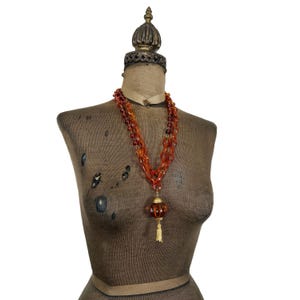 May include: A multi-strand necklace featuring translucent amber-colored beads and oval links. The necklace includes a central spherical pendant with gold-toned metal accents and a small hanging tassel, displayed on a fabric-covered mannequin torso.