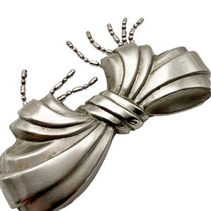May include: A silver bow-shaped brooch with a textured surface and a chain detail. The chain is made of small, round beads.