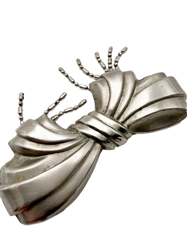 May include: A silver bow-shaped brooch with a textured surface and a chain detail. The chain is made of small, round beads.