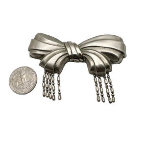 May include: A silver bow brooch with a dangling chain of beads. The brooch is about the size of a US quarter.