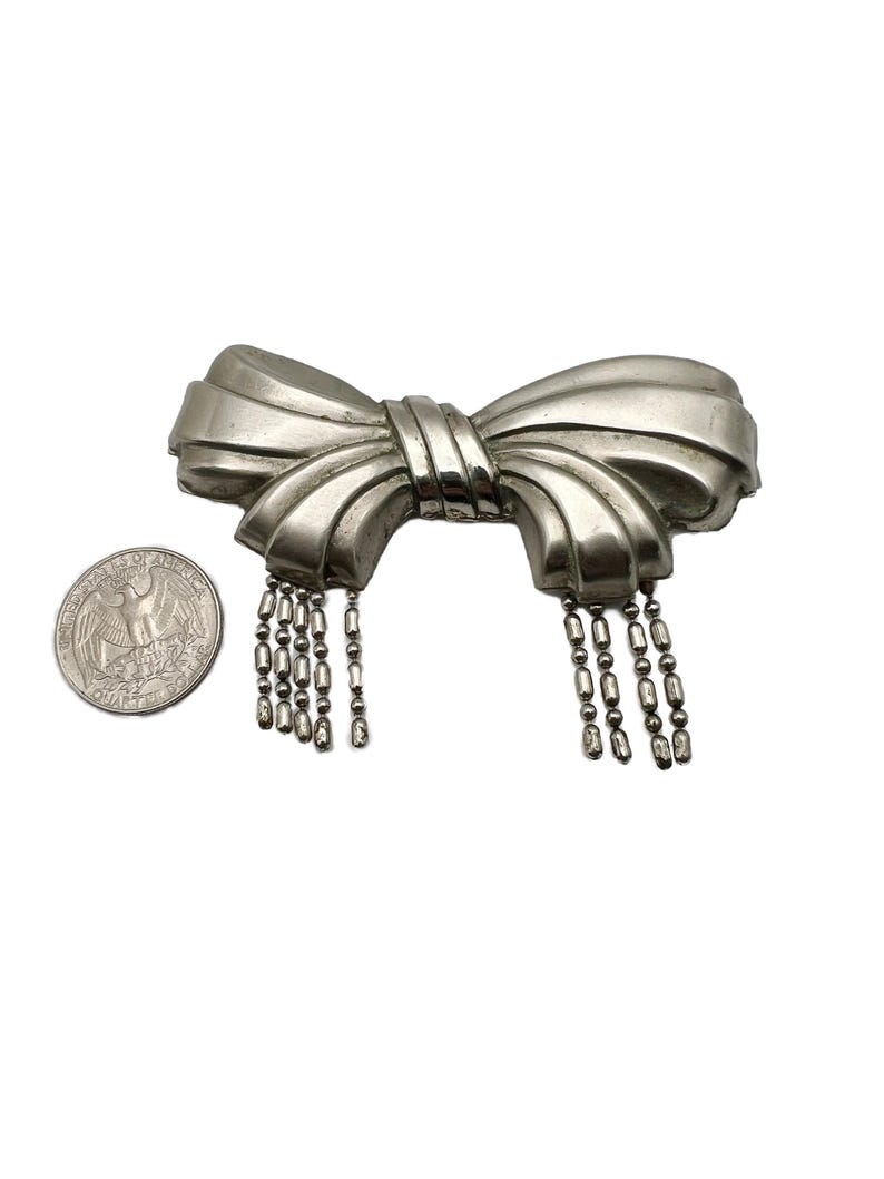 May include: A silver bow brooch with a dangling chain of beads. The brooch is about the size of a US quarter.
