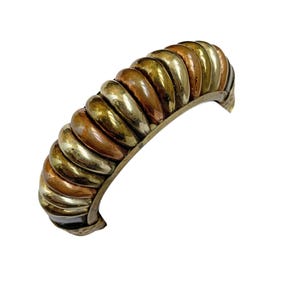 Puede incluir: Un brazalete de oro, cobre y plata con un diseño texturizado y redondeado. El brazalete está hecho de múltiples bandas de metal que están entrelazadas.