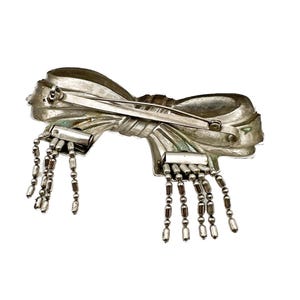 May include: A silver bow brooch with dangling chains of beads. The bow is detailed with a textured surface and a pin on the back.