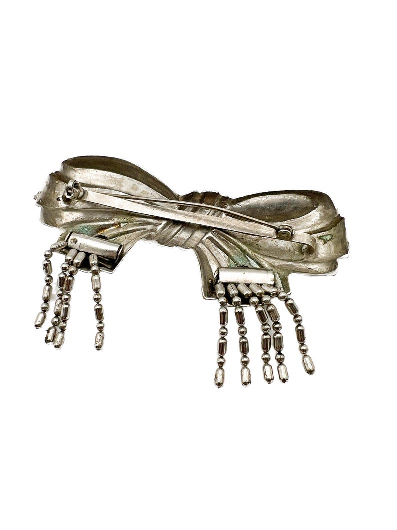 May include: A silver bow brooch with dangling chains of beads. The bow is detailed with a textured surface and a pin on the back.