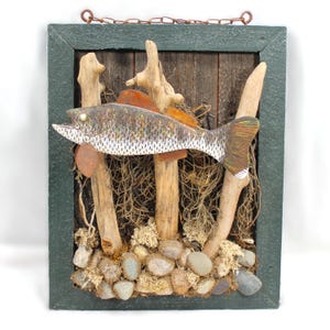 May include: A wall-mounted art piece featuring a carved wooden fish mounted on driftwood, set against a dark wooden background within a dark green frame. The base is decorated with small grey stones and dried moss, with a metal chain for hanging.