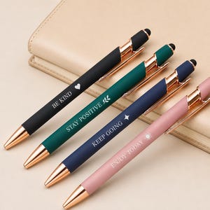 Könnte beinhalten: Vier Stifte mit roségoldfarbenen Akzenten und schwarzen Touch-Spitzen. Die Stifte sind schwarz, grün, marineblau und rosa, jeweils mit einem inspirierenden Satz in Weiß: "BE KIND", "STAY POSITIVE", "KEEP GOING" und "ENJOY TODAY".