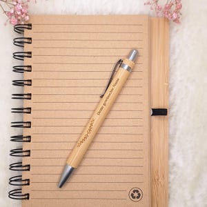 Może przedstawiać: Brązowy notes ze spiralnym oprawą i liniowanymi stronami. Notes jest wykonany z bambusa i ma symbol recyklingu na okładce. Drewniany długopis ze srebrnym końc&oacute;wką leży na notesie. Długopis jest grawerowany tekstem "Deine gew&uuml;nschte Name" i wzorem kwiatowym.
