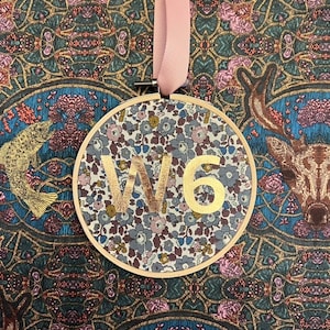 May include: A round, wooden ornament with a floral pattern and the text "W16" in gold lettering. The ornament is hanging from a pink ribbon.