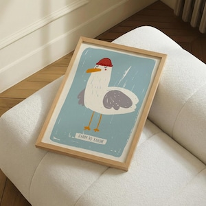 Puede incluir: Un cuadro enmarcado con una ilustración de una gaviota de dibujos animados con un gorro rojo. La gaviota tiene alas grises y patas amarillas, sobre un fondo azul claro. El texto "READY TO EXPLORE" está en la parte inferior. El marco es de madera clara.