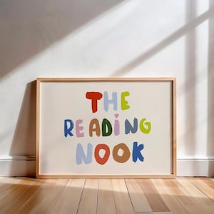 Puede incluir: Una lámina decorativa rectangular en un marco de madera clara con el texto THE READING NOOK en letras multicolores de estilo pintado a mano. Las letras están representadas en tonos rojo, azul, verde, rosa, marrón y morado claro sobre fondo crema.