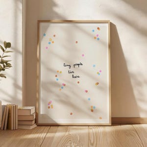 Puede incluir: Una impresión artística enmarcada con las palabras "tiny people live here" en escritura negra. La impresión tiene un fondo crema con puntos coloridos dispersos en tonos de rosa, naranja, azul y amarillo. El marco es de color madera clara.