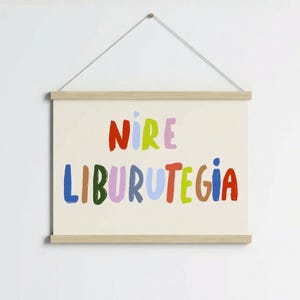 Op de afbeelding: Een rechthoekige wandhanger met een lichtcrème achtergrond en een houten frame met een ophangkoord. De tekst NIRE LIBURUTEGIA is gedrukt in grote, veelkleurige, handgetekende letters in tinten rood, groen, blauw en roze.