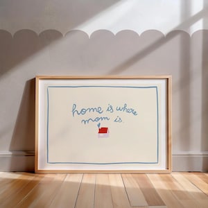 Puede incluir: Una impresión artística enmarcada con el texto "home is where mom is." La obra presenta un fondo beige claro con un borde azul y una pequeña ilustración de una casa con un corazón. El marco es de madera clara.