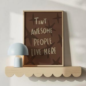 Puede incluir: Un estampado enmarcado con el texto "Tiny Awesome People Live Here" e ilustraciones de estrellas. Una lámpara en forma de hongo azul claro se encuentra en una estantería de madera con volantes. El estampado tiene un fondo marrón.