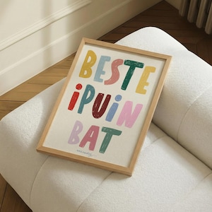 Puede incluir: Una impresión artística enmarcada con las palabras "BESTE !PUIN BAT" en letras coloridas y divertidas. La obra de arte está sobre un fondo color crema y enmarcada en madera clara. La impresión descansa sobre una superficie blanca y texturizada.