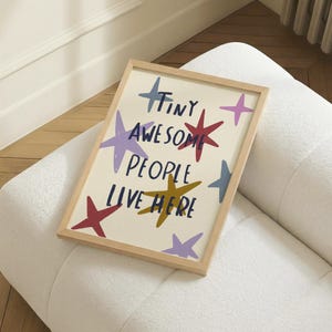 Puede incluir: Un cuadro enmarcado con la frase "TINY AWESOME PEOPLE LIVE HERE" en azul marino, con ilustraciones de estrellas coloridas. Las estrellas son de color rojo, morado, amarillo y azul. El marco es de madera clara.