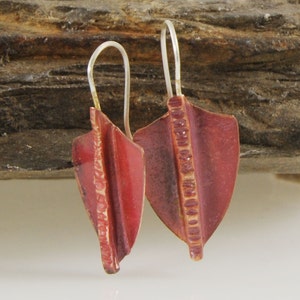 May include: A pair of shield-shaped earrings in a deep red hue, featuring a textured vertical stripe. The ear wires are silver. The earrings are displayed against a natural wood background. The earrings are approximately 3 cm in length.