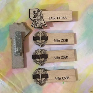 Puede incluir: Tres etiquetas de nombre de madera con texto y diseños grabados. La etiqueta superior dice "2ABCT FRSA". Las etiquetas del medio y la inferior dicen "541st CSSB". Cada etiqueta presenta un diseño diferente, incluyendo un león, una cruz y una pancarta.