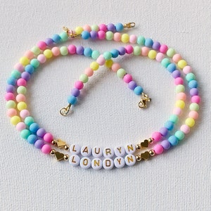 May include: Two layered pastel rainbow beaded necklaces with gold heart charms and white letter beads spelling out the names "Lauryn" and "Londyn".