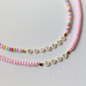 May include: Two beaded necklaces featuring small, faceted, multi-colored beads and solid pink beads. Each necklace includes white circular beads with gold lettering spelling out the names BRINLEY and EVIE, accented by small gold heart-shaped beads.
