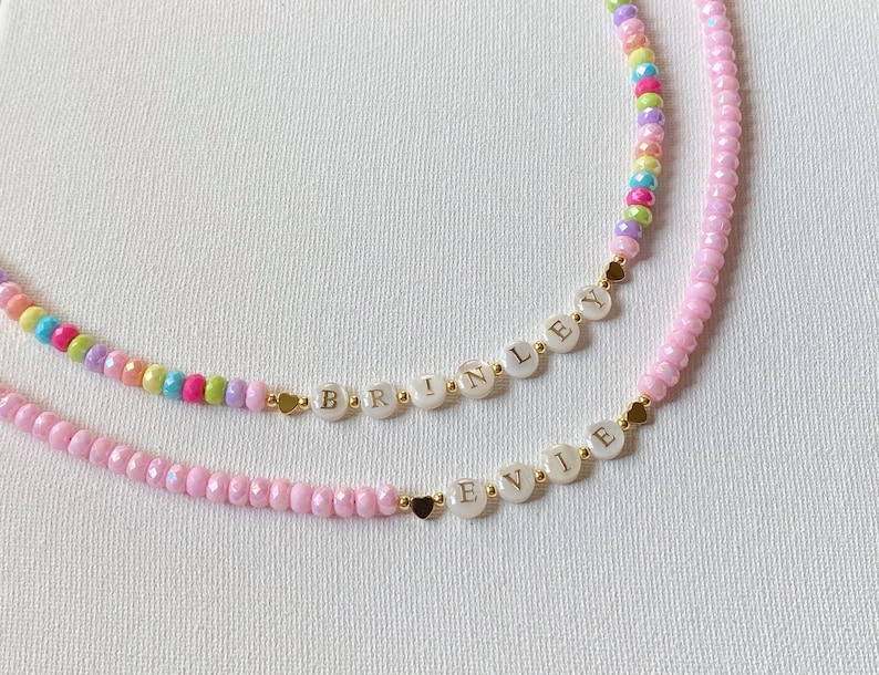 May include: Two beaded necklaces featuring small, faceted, multi-colored beads and solid pink beads. Each necklace includes white circular beads with gold lettering spelling out the names BRINLEY and EVIE, accented by small gold heart-shaped beads.