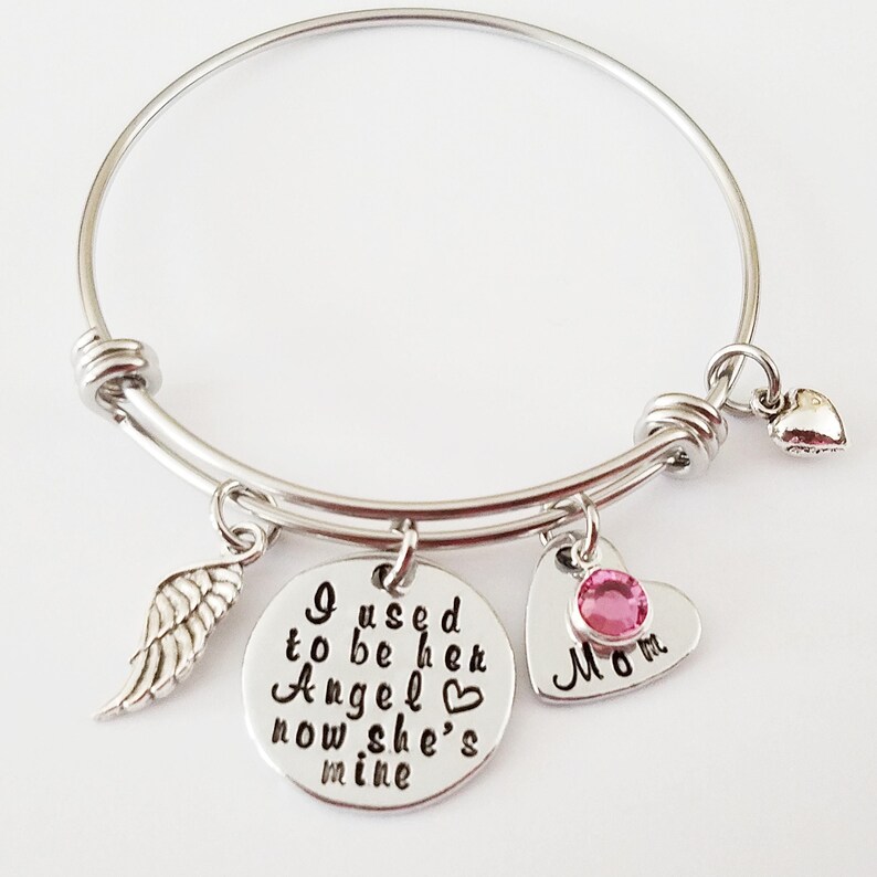 May include: A silver bangle bracelet with a charm that reads "I used to be her Angel, now she's mine" with a small heart, a silver wing charm, and a heart charm with a pink rhinestone that reads "Mom".