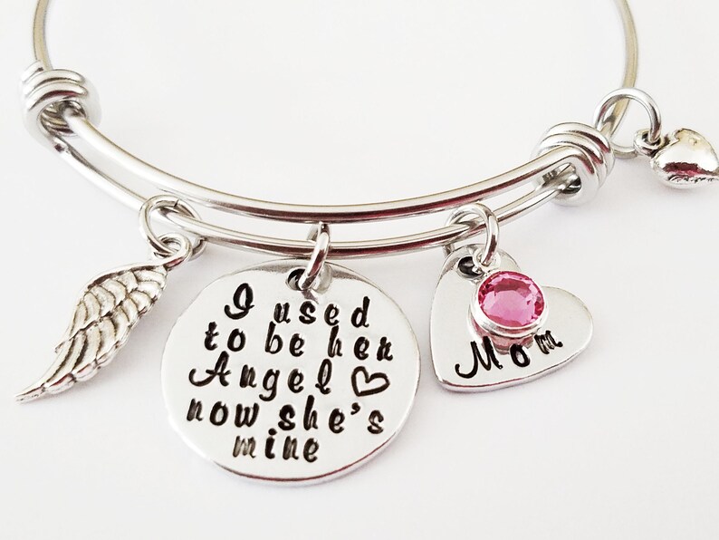 May include: Silver bangle bracelet with three charms: a wing, a heart with a pink stone that says "Mom", and a round charm that says "I used to be her Angel now she's mine".