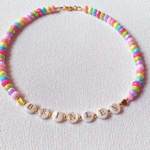 May include: A beaded necklace featuring a repeating pattern of small, multi-colored faceted beads in shades of pink, yellow, blue, green, and purple. The center includes white circular beads with the gold-colored letters spelling BRINLEY.