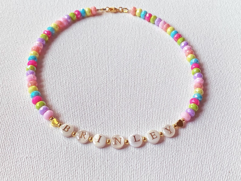 May include: A beaded necklace featuring a repeating pattern of small, multi-colored faceted beads in shades of pink, yellow, blue, green, and purple. The center includes white circular beads with the gold-colored letters spelling BRINLEY.