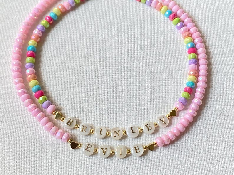 May include: Two beaded necklaces featuring pink, yellow, blue, green, and purple faceted beads. One necklace displays the name BRINLEY and the other displays the name EVIE on white circular beads with gold lettering, accented by small gold heart beads.