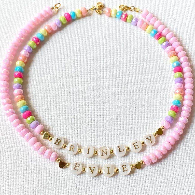 May include: Two beaded necklaces featuring pastel-colored faceted beads and gold-tone metal accents. One necklace displays the name BRINLEY and the other displays the name EVIE using white circular letter beads with gold lettering and small heart charms.