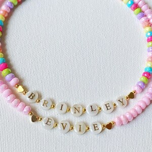 May include: Two beaded necklaces featuring faceted light pink beads and a strand of multicolored beads. One necklace displays the name BRINLEY and the other displays the name EVIE on white circular beads with gold lettering and small gold heart accents.