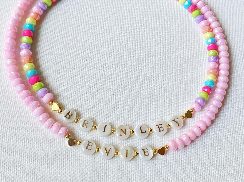 May include: Two beaded necklaces featuring faceted light pink beads and a strand of multicolored beads. One necklace displays the name BRINLEY and the other displays the name EVIE on white circular beads with gold lettering and small gold heart accents.