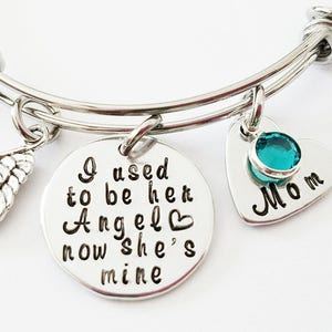 May include: A silver bangle bracelet with three charms. The charms are a small angel wing, a heart-shaped charm with the text "I used to be her Angel now she's mine" and a heart-shaped charm with a turquoise stone and the text "Mom".