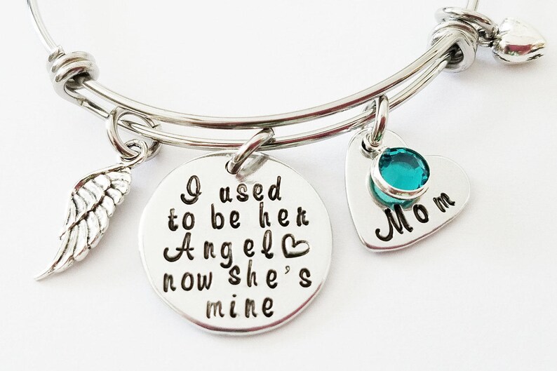 May include: A silver bangle bracelet with three charms. The charms are a small angel wing, a heart-shaped charm with the text "I used to be her Angel now she's mine" and a heart-shaped charm with a turquoise stone and the text "Mom".