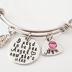 May include: Silver bangle bracelet with charms. The charms include a wing, a heart with a pink rhinestone, and a round charm with the text "I used to be her Angel now she's mine".