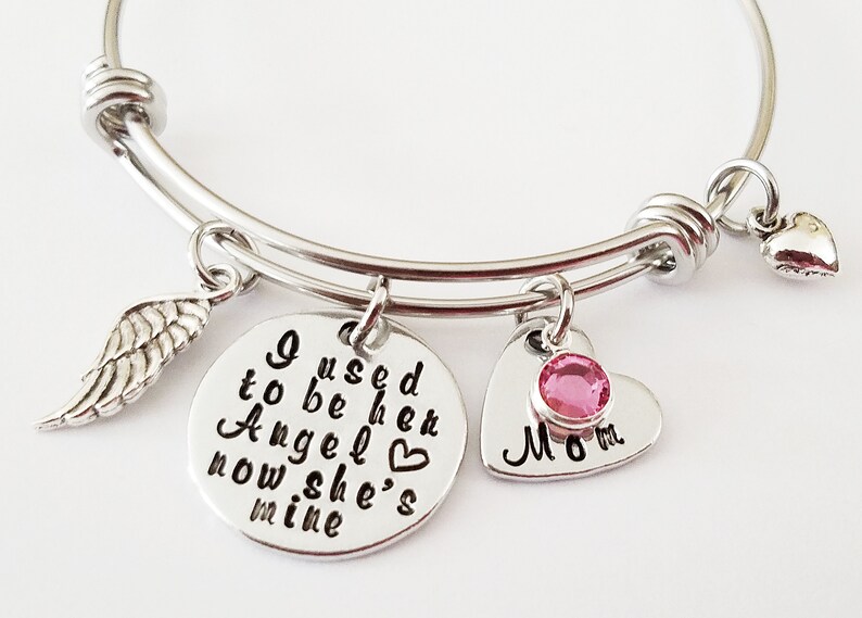 May include: Silver bangle bracelet with charms. The charms include a wing, a heart with a pink rhinestone, and a round charm with the text "I used to be her Angel now she's mine".