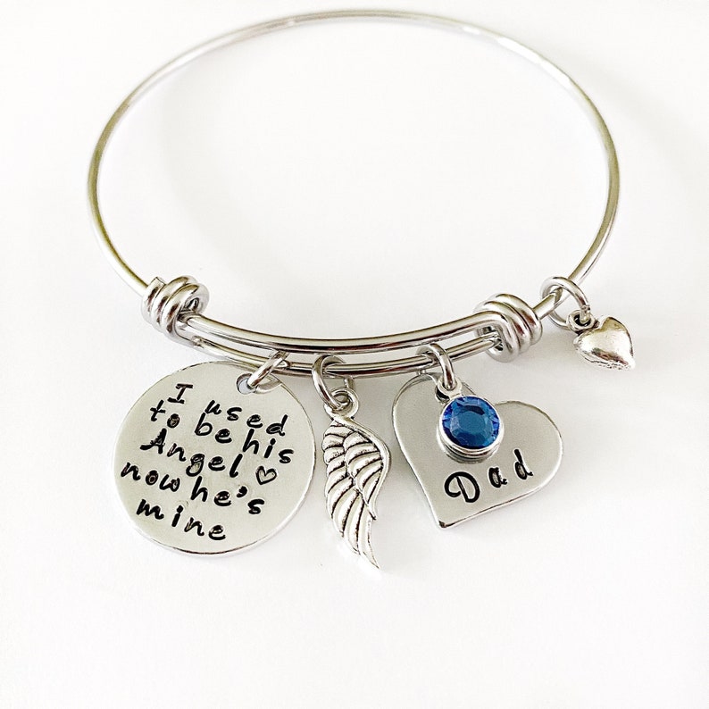 May include: Silver bangle bracelet with charms. The charms include a round charm with the text "I used to be his Angel now he's mine", a heart charm with a blue rhinestone and the text "Dad", and a silver angel wing charm.