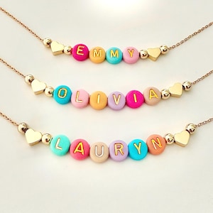 May include: Three gold-colored necklaces with personalized name beads. Each necklace features a different name spelled out with colorful round beads and gold-colored letters. Gold heart and ball accents are also present.
