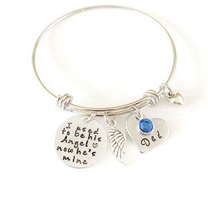May include: Silver bangle bracelet with charms. The charms include a round charm with the text "I used to be his Angel now he's mine" and a heart charm with a blue crystal and the text "Dad".