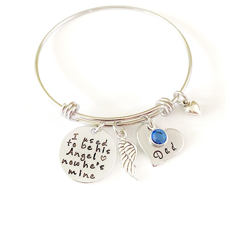 May include: Silver bangle bracelet with charms. The charms include a round charm with the text "I used to be his Angel now he's mine" and a heart charm with a blue crystal and the text "Dad".